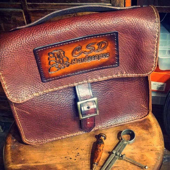 Reddish brown messenger bag with buckle clasp and custom tooled logo for CSD Hardscaped on top flap.