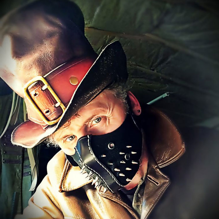 Don wearing a brown leather top hat with red brown leather band with buckle, and a black stiff leather mask with spikes.