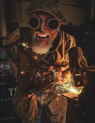 Don Dodson in a steampunk ironwork costume, looking at the camera with mouth open "yelling", grinding an mallet.